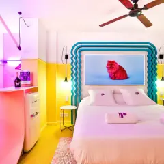 Pokój w stylu memphis z łóżkiem, lodówką Smeg i obrazem czerwonego kota. Kolorowe ściany i meble tworzą radosną atmosferę.